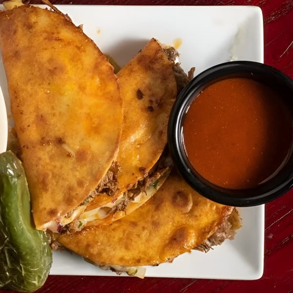Three golden-brown Birrieria Esquivel birria tacos with a side of red dipping sauce and a green pepper.